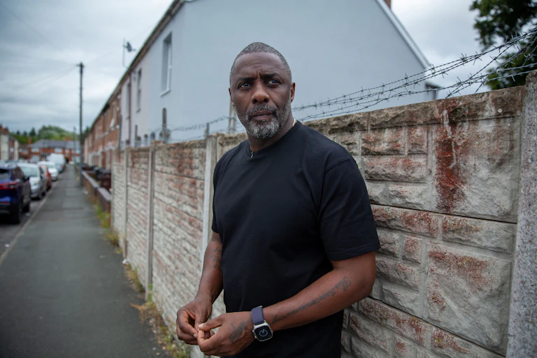 Idris Elba stands against suburban wall topped with barbed wire