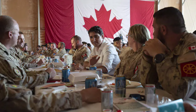 A dark-haired man sits among uniformed soldiers with the Canadian flag in the background.