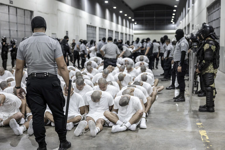 Men with shaved heads and in white shirts and shorts sitting on a prison floor with guards around them.