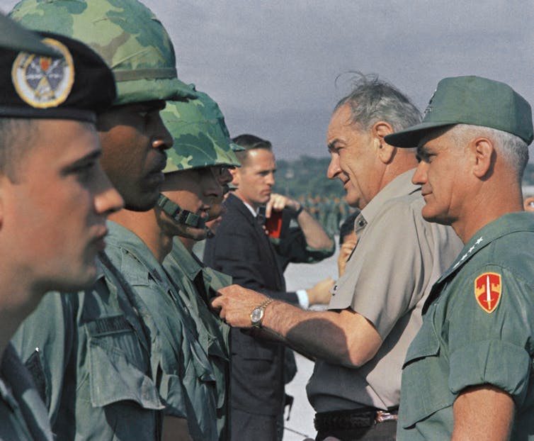A man in civilian clothes and a man in a military uniform stand facing a row of men in military uniforms.
