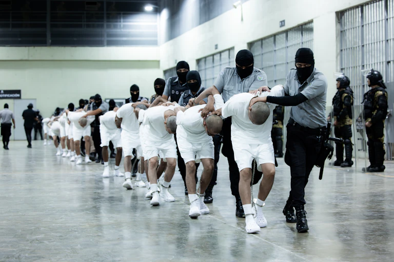 Men in white shirts and shorts are bent double, being led by arimed men in balaclavas