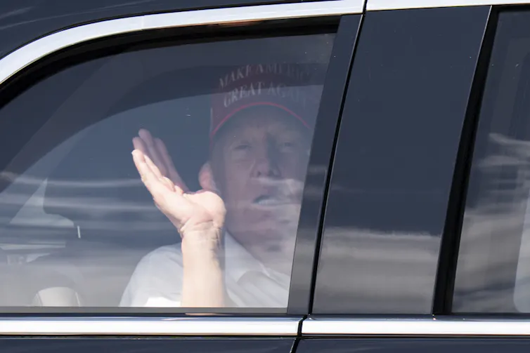 An elderly, exhausted-looking man waves from the back of a limousine. He's wearing a red ball cap.