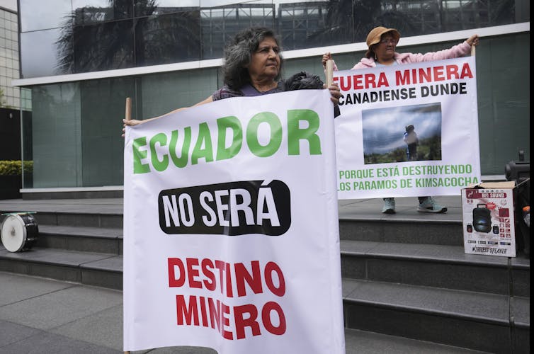 Two Ecuadorian people hold up signs protesting against mining operations in the country. Their signs say: 'Ecuador will not be a mining destiny,' and 'Get out Dunde mining company, because you are destroying Los Paramos of Kimsachocha'