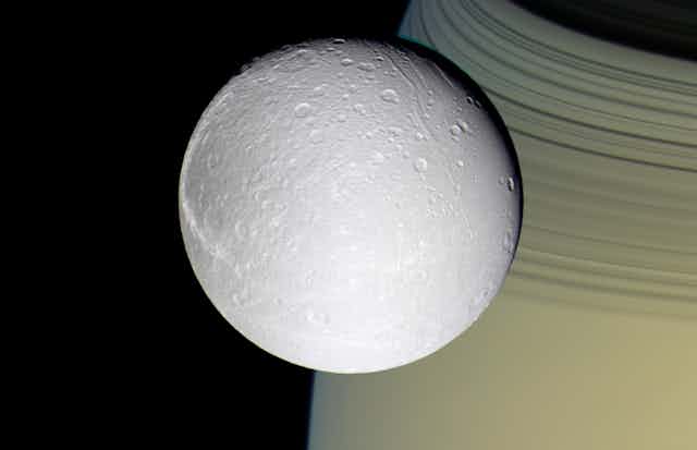 Foto de uma lua gelada com um planeta amarelado e anéis atrás,