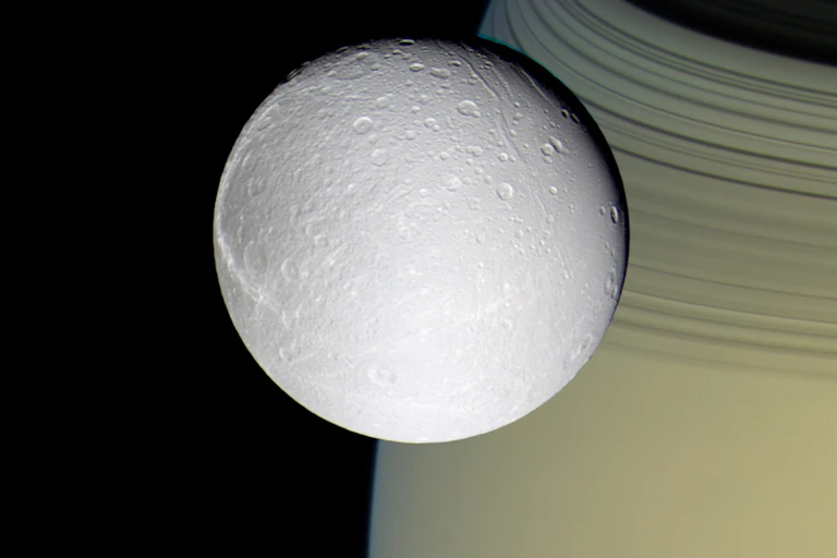 Foto de uma lua gelada com um planeta amarelado e anéis atrás,