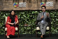 A white man with dark hair in a grey suit speaks into a microphone while seated. Beside him sits an Asian woman in a red suit.