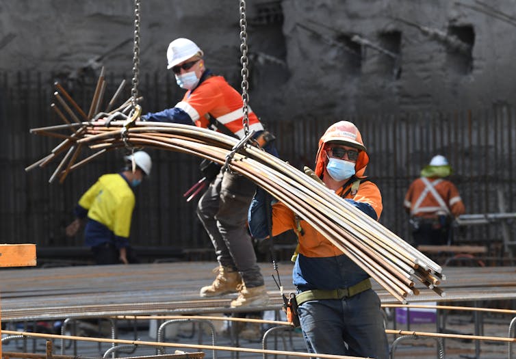 Two workers, wearing hard hats and hi-viz clothing, manoeuvring large rods on a building site.