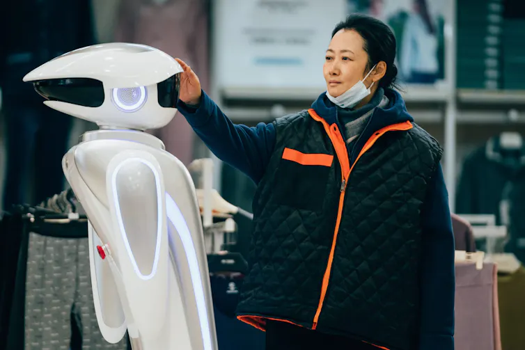A woman has a face mask under her chin. She strokes a robot.