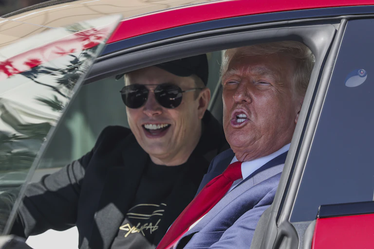 A man dressed in black wearing sunglasses smiles next to an older man with heavy facial bronzer and white-blond hair.