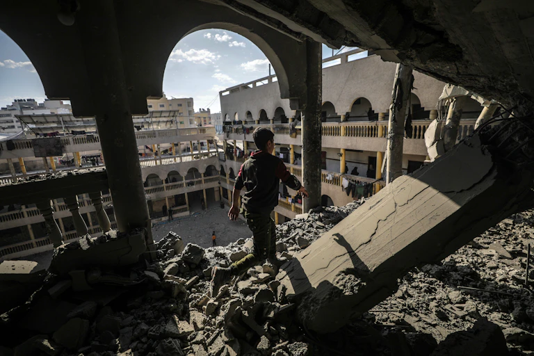 A boy stands in an upper floor of a building that has been bombed looking out at the rubble of other buildings.