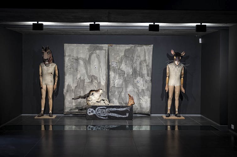 Two human figures with animal heads stand on either side of a box with a drawing of a skeleton and a buck head on top of it, against a backdrop of a childlike painting.