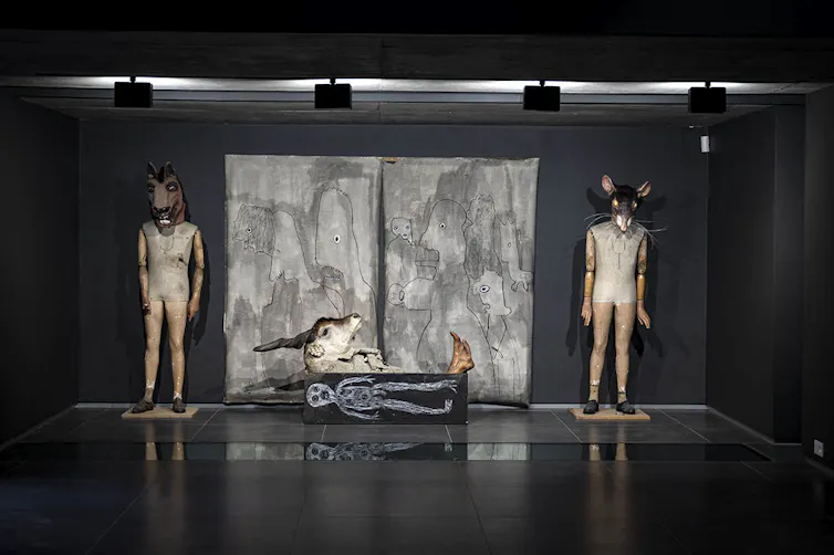 Two human figures with animal heads stand on either side of a box with a drawing of a skeleton and a buck head on top of it, against a backdrop of a childlike painting.