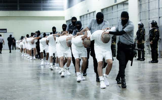 Men in white shirts and shorts are bent double, being led by arimed men in balaclavas