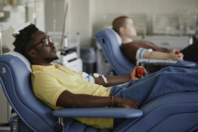 Two people lie on reclining seats, with cuffs around their upper arms and needles inserted at their elbows