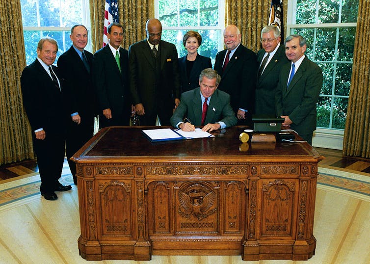 Man seated at desk signs piece of paper while surrounded by seven men and one woman.