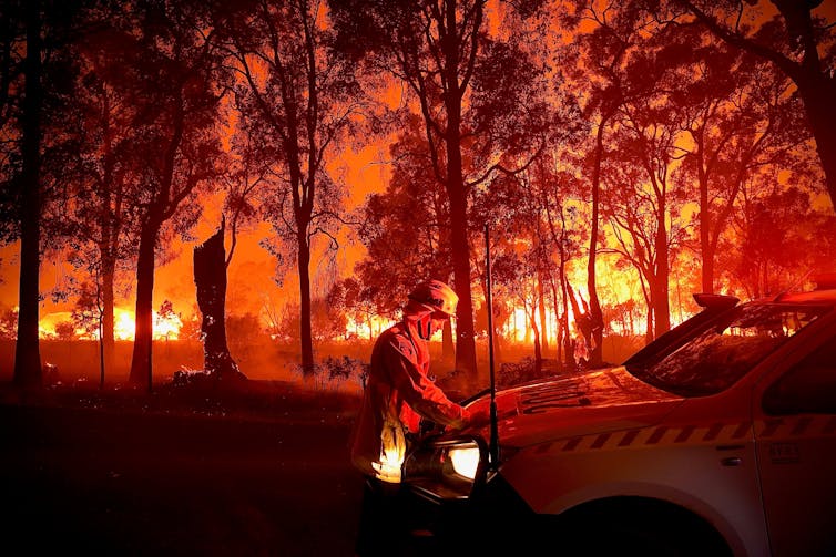 A fireman wearing protective gear standing in front off a vehicle parked next to trees which are threatened by an approaching bush fire