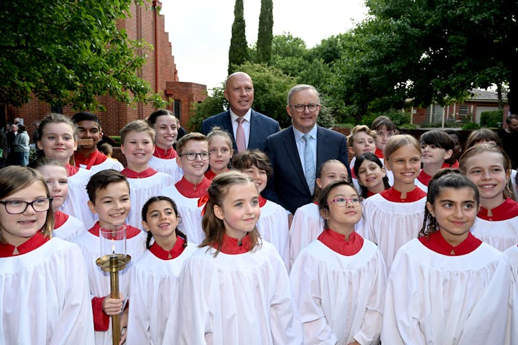Anthony Albanese and Peter Dutton standing behind more than than a dozen children who are wearing white smocks with red collars