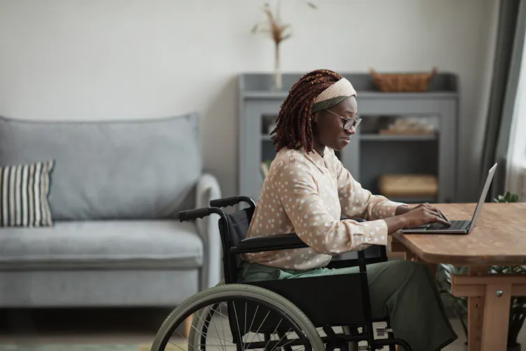 A young woman in a wheelchair sitting at a desk at home, working on a laptop
