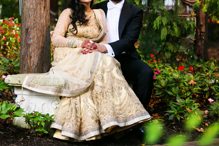 Woman with south Asian style wedding dress sat holding hands with man wearing white tie morning suit.