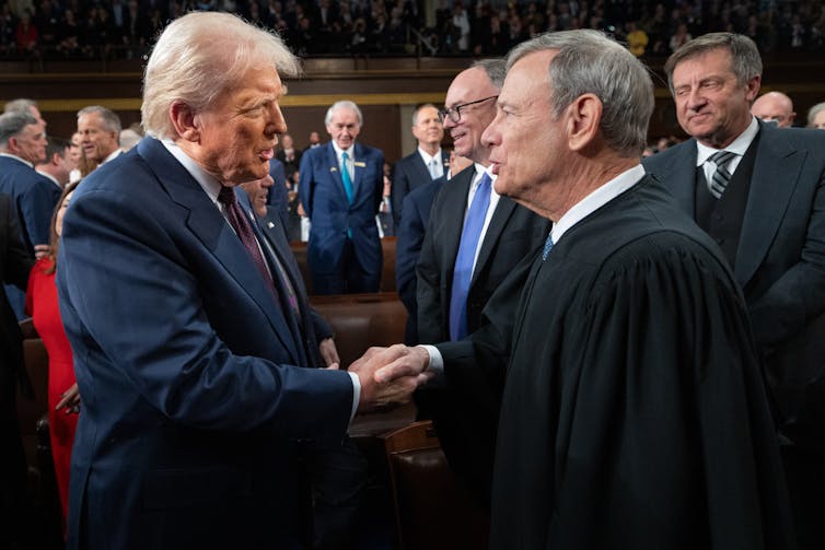 Two men, one wearing a dark suit and with white hair, and the other with a black robe, shake hands.