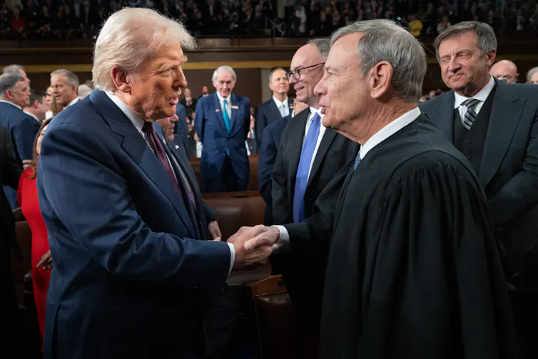 Two men, one wearing a dark suit and with white hair, and the other with a black robe, shake hands.