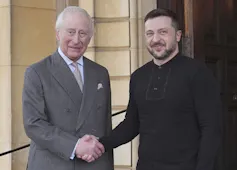 An older man in a suit shakes hands with a dark-haired bearded man in dark clothing.