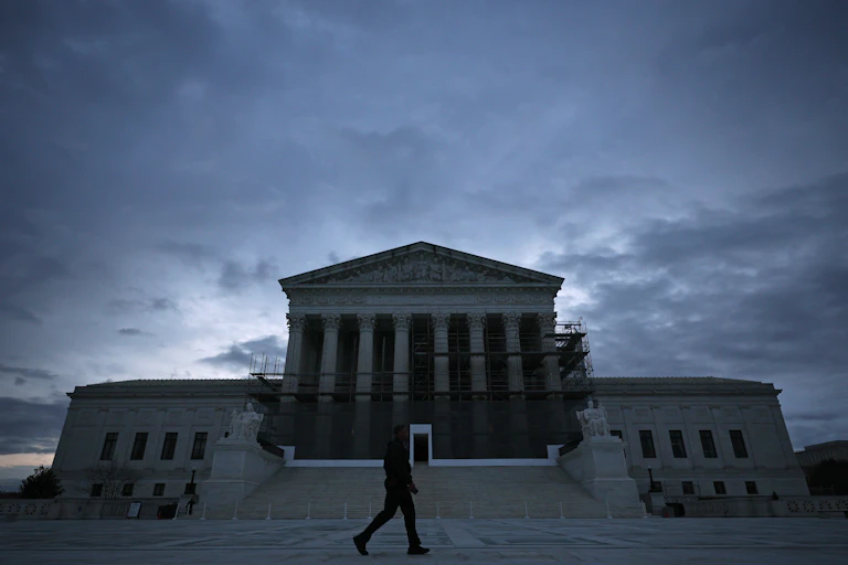 A person walks past a tall building with columns with a gray and blue sky.