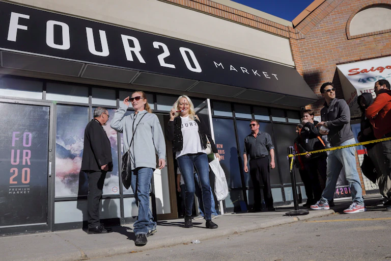 Two women walk out of a cannabis store in Canada. A line of people wait beside them to get in.