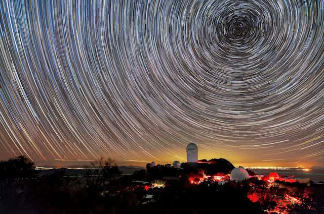 Timelapse photo showing star trails in the night sky above a large telescope building.