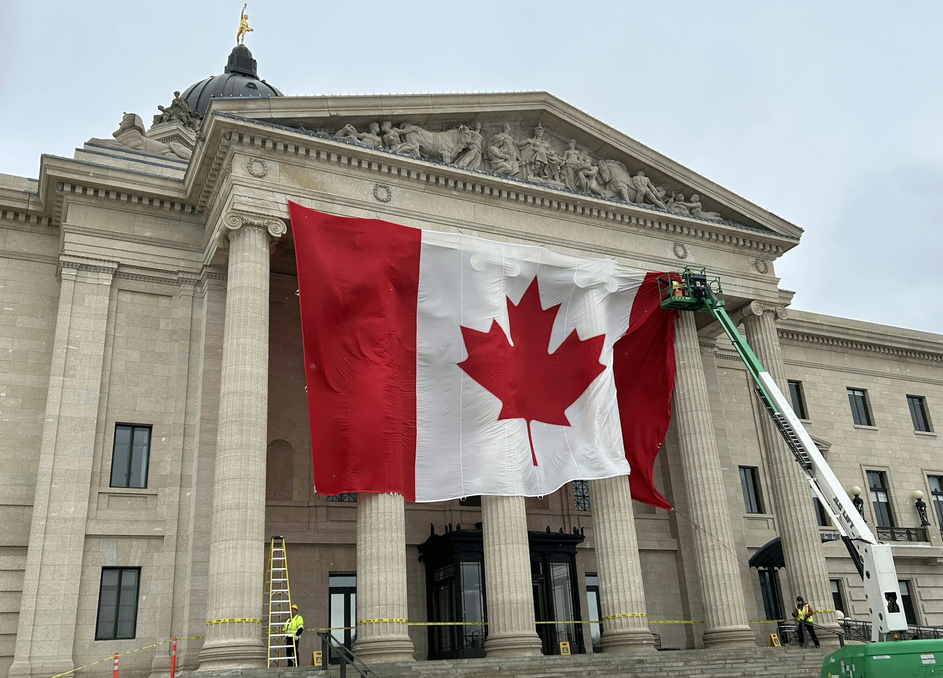 A large Canadian flag is seen being mounted by a crane in front of a tall stone building with columns.