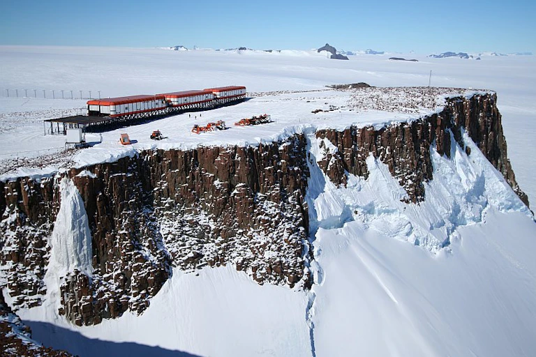 A large research base, consisting of three white and red modules, located on the side of a steep cliff, surrounded by snow and ice.
