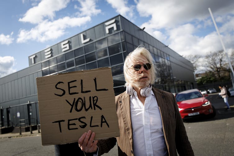 A white-bearded man in sunglasses holds a sign, 'Sell your Tesla', in front of a Tesla building.