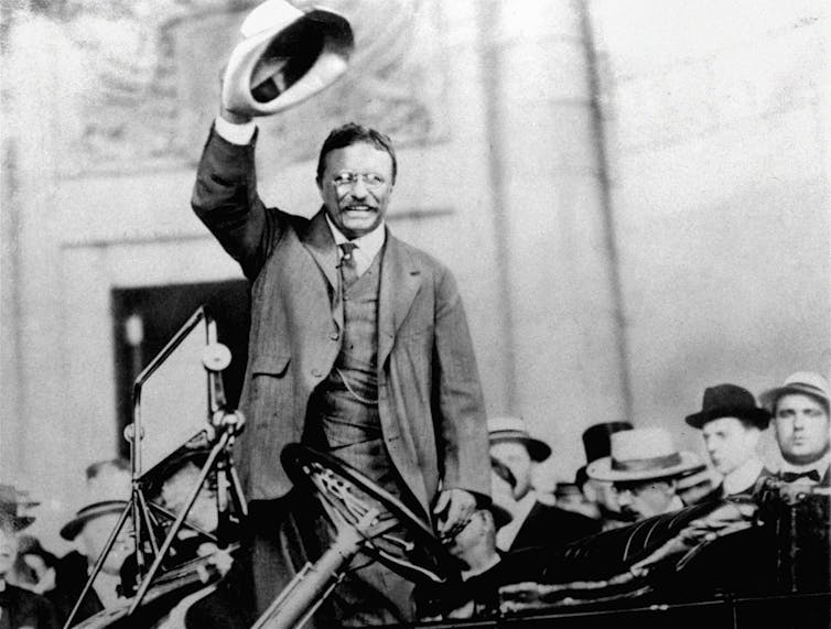 A man in a suit in a car amidst a crowd, waving his hat high