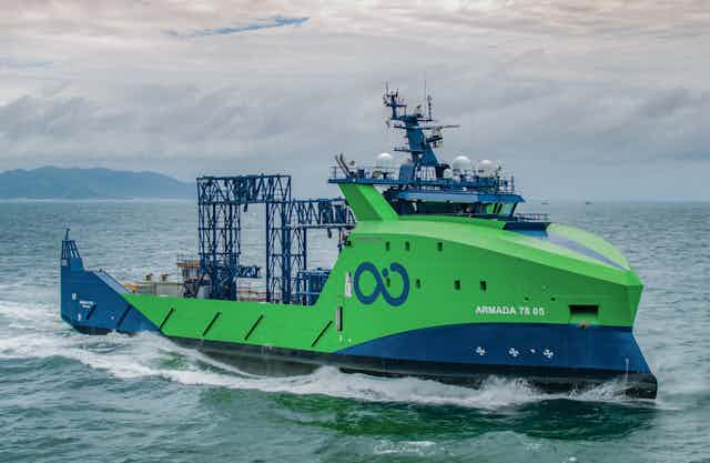 A blue and green ship powering through ocean waves under cloudy sky.