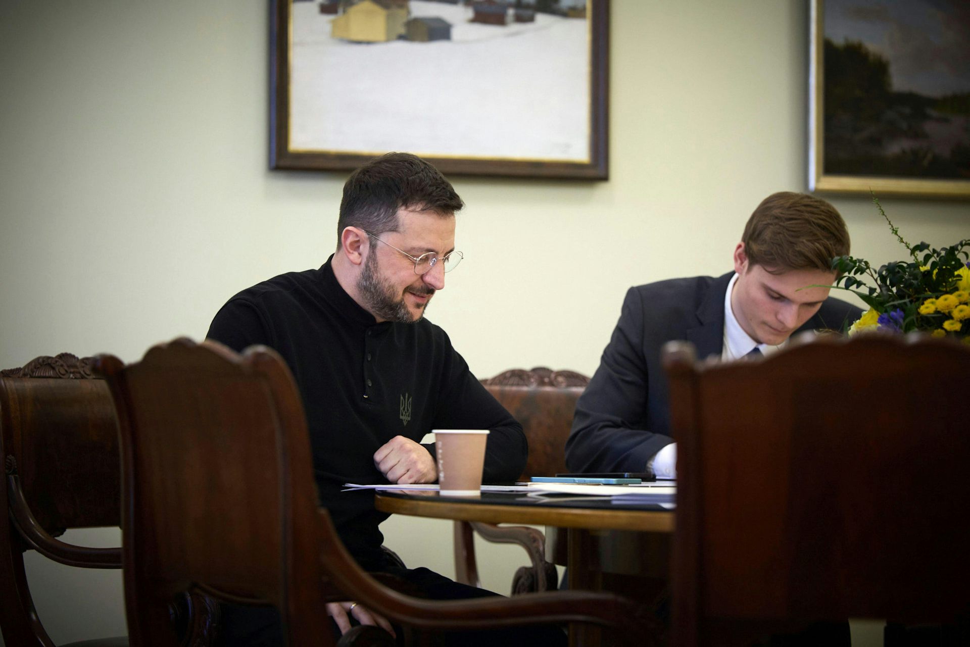 Ukrainian president, Volodymyr Zelensky, sits at a table talking on the telephone while an aide listens in.