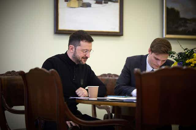 Ukrainian president, Volodymyr Zelensky, sits at a table talking on the telephone while an aide listens in.