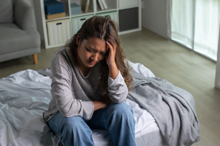 A young woman sitting on a bed in her bedroom, head in her hands, looking anxious