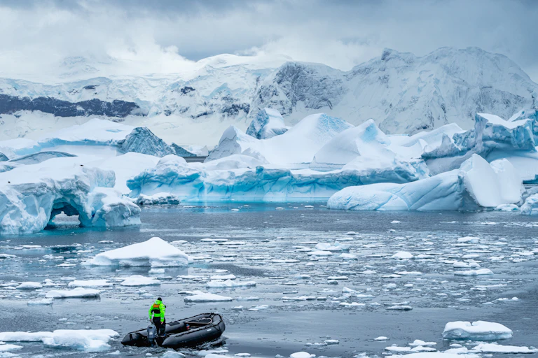 A small boat drives through melted ice with a backdrop of ice bergs and shelves.