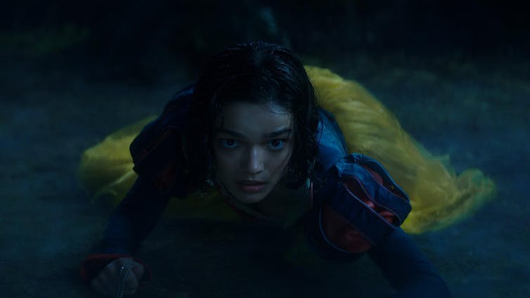 Una chica mojada, en el suelo, con un vestido azul y amarillo, mira a cámara.