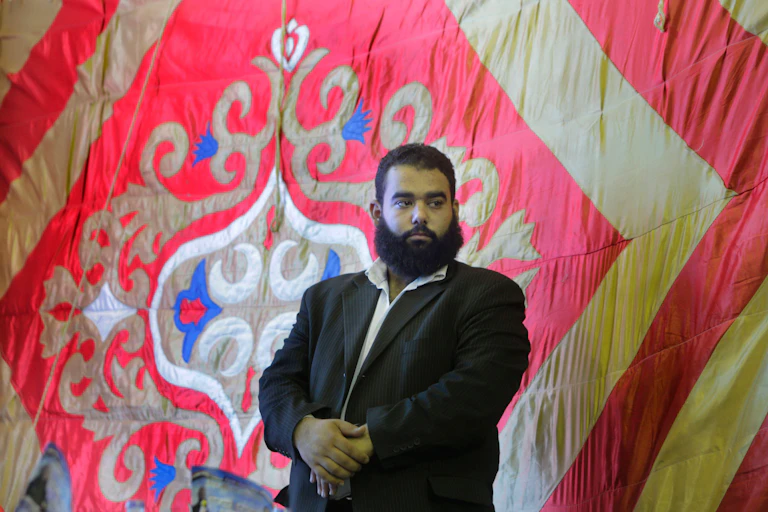 A man with a beard stands in front of a wall hanging displaying artistic calligraphy.