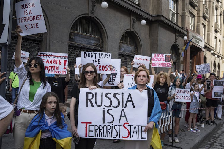 Protesters line a city street. One sign reads Russia is a Terrorist State.