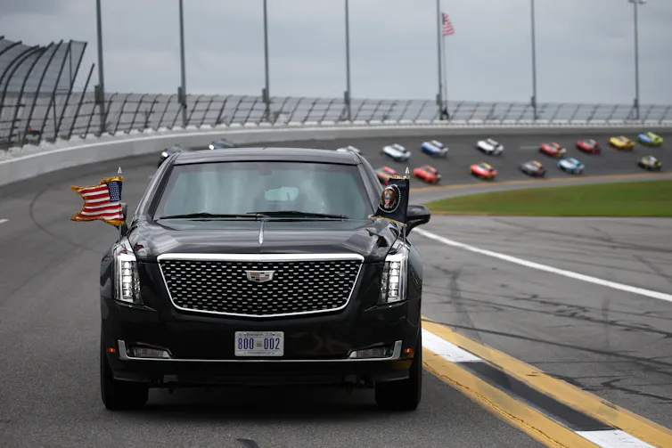 A black car with American flags drives around a racetrack, with race cars behind it.
