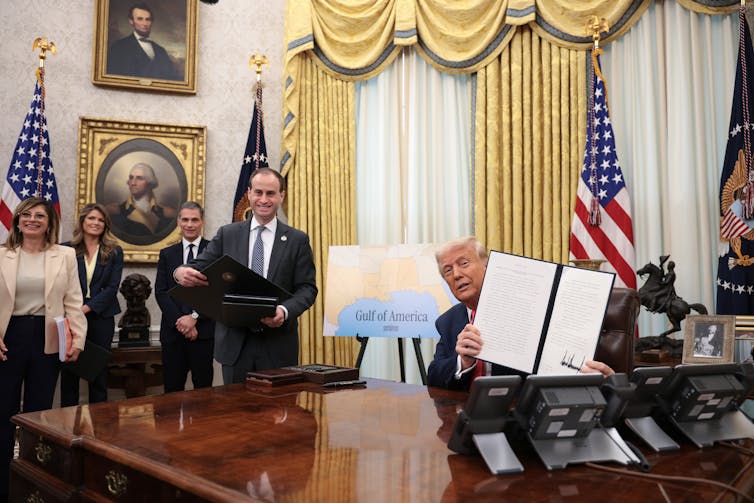 A man with white hair is seated at a table and holds up a folder showing white pages and a signature. He sits in an office with other people and American flags.