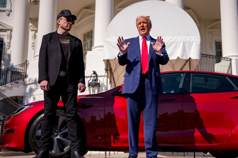 A man with a blue suit and a red tie stands next to a man dressed all in black in front of a red sports car.