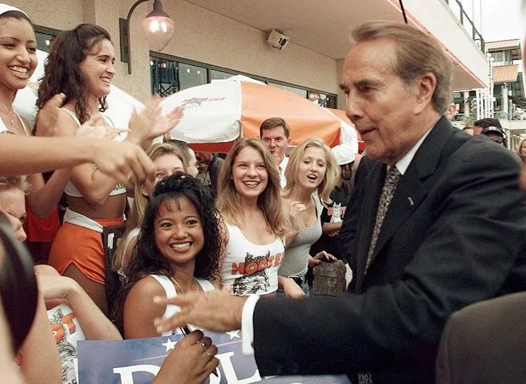 Older man in suit greets crowd of young women dressed in white and orange.
