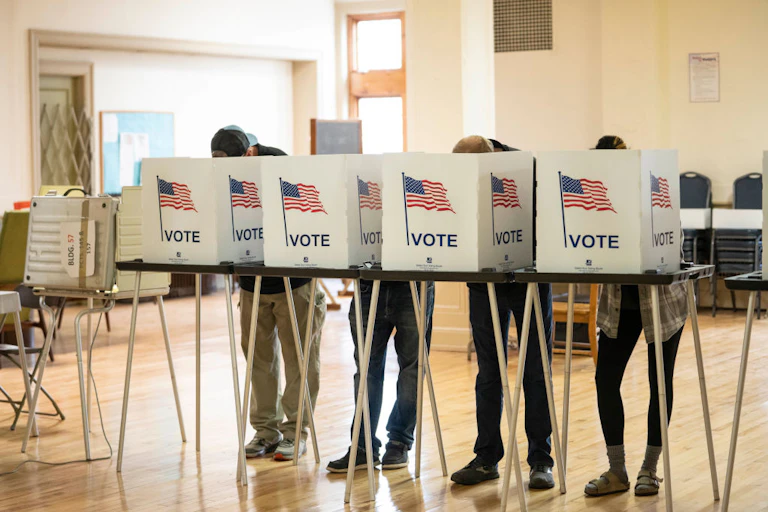 Four people bend over a tall folding table, their faces hidden by folders that say 'vote' with American flags printed on them.