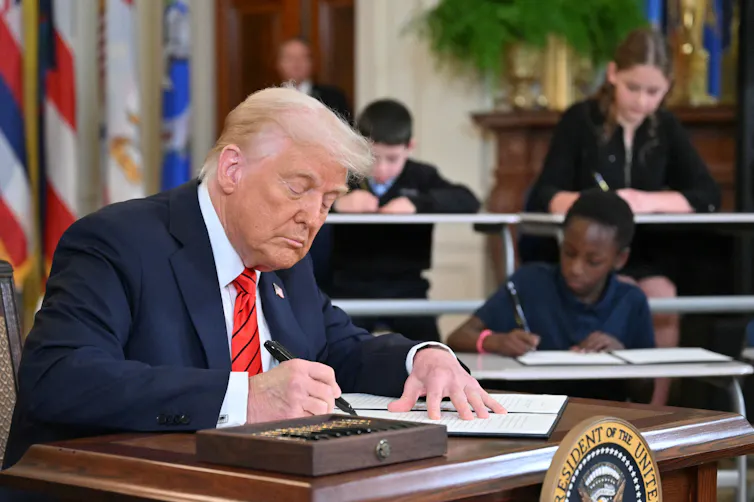 A man wearing a blue suit and a red tie writes on a piece of paper, with several students sitting at desks behind him.