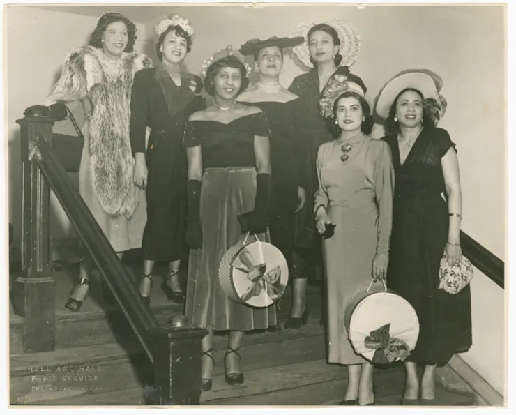 Sepia-toned photo of a a group of seven fashionable women wearing hats pose together on stairs