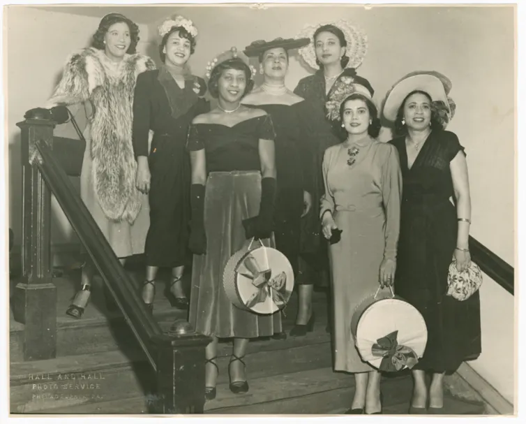 Sepia-toned photo of a a group of seven fashionable women wearing hats pose together on stairs