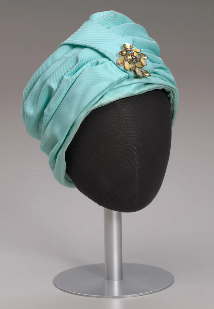 Head mannequin wears turquoise turban-style hat with gold-colored gem brooch
